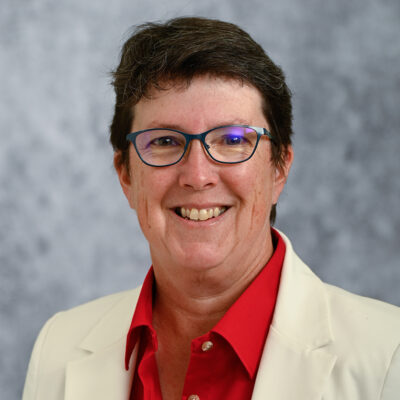 Professional photo of Evelyn Brown wearing glasses, a red shirt and tan coat in front of a grey background.