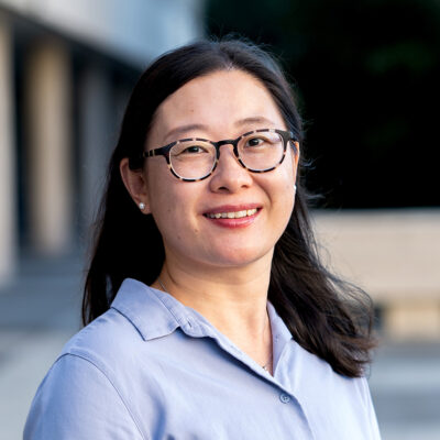 Zhina Shen headshot outdoors, blue button down and glasses