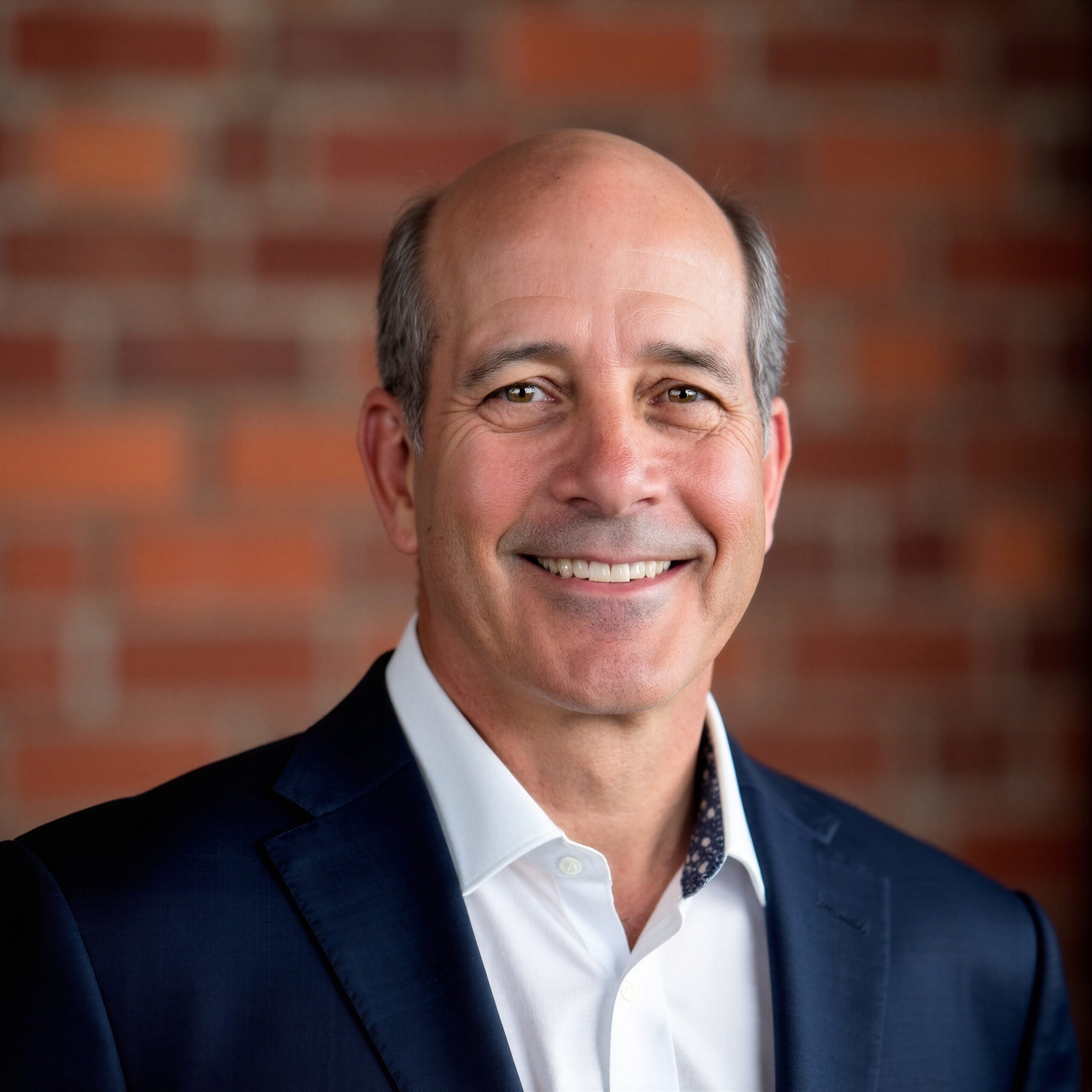 Close-up headshot of an adult person wearing a dark blazer and white collared shirt, facing the camera against a softly blurred brick wall background.
