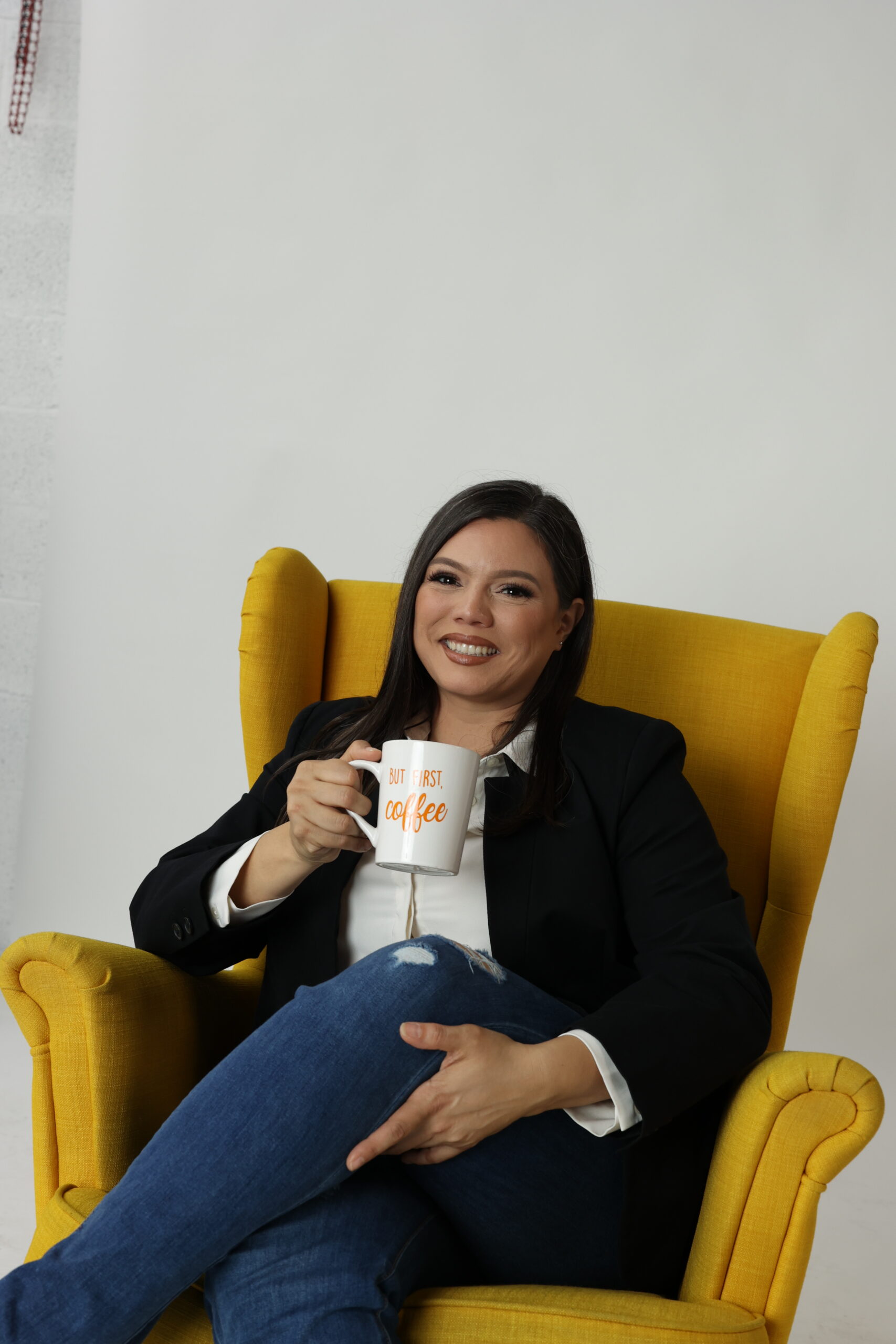 A casual photo of Jacqueline Pena sitting in a yellow chair holding a mug that says "But first, coffee".