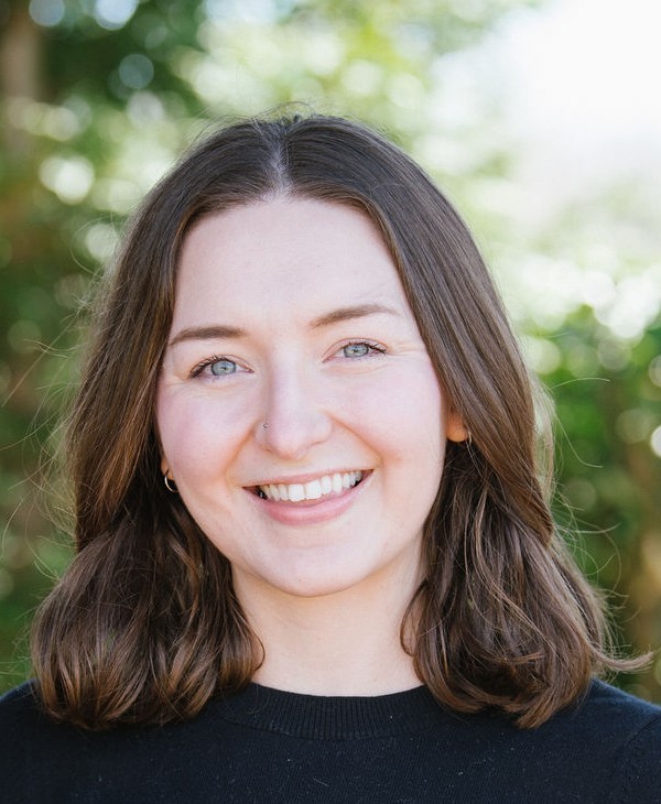 Close-up outdoor portrait of Jen Gruber with shoulder-length brown hair wearing a black top, facing the camera, with a softly blurred green foliage background.