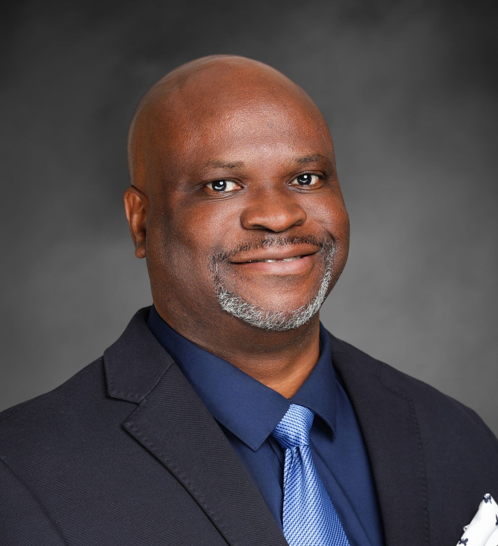 Professional studio headshot of Adeyemo Adetogun Ph.D. wearing a dark suit jacket, navy shirt, and light blue tie, with a patterned pocket square. The portrait is set against a neutral gray background and framed from the chest up.