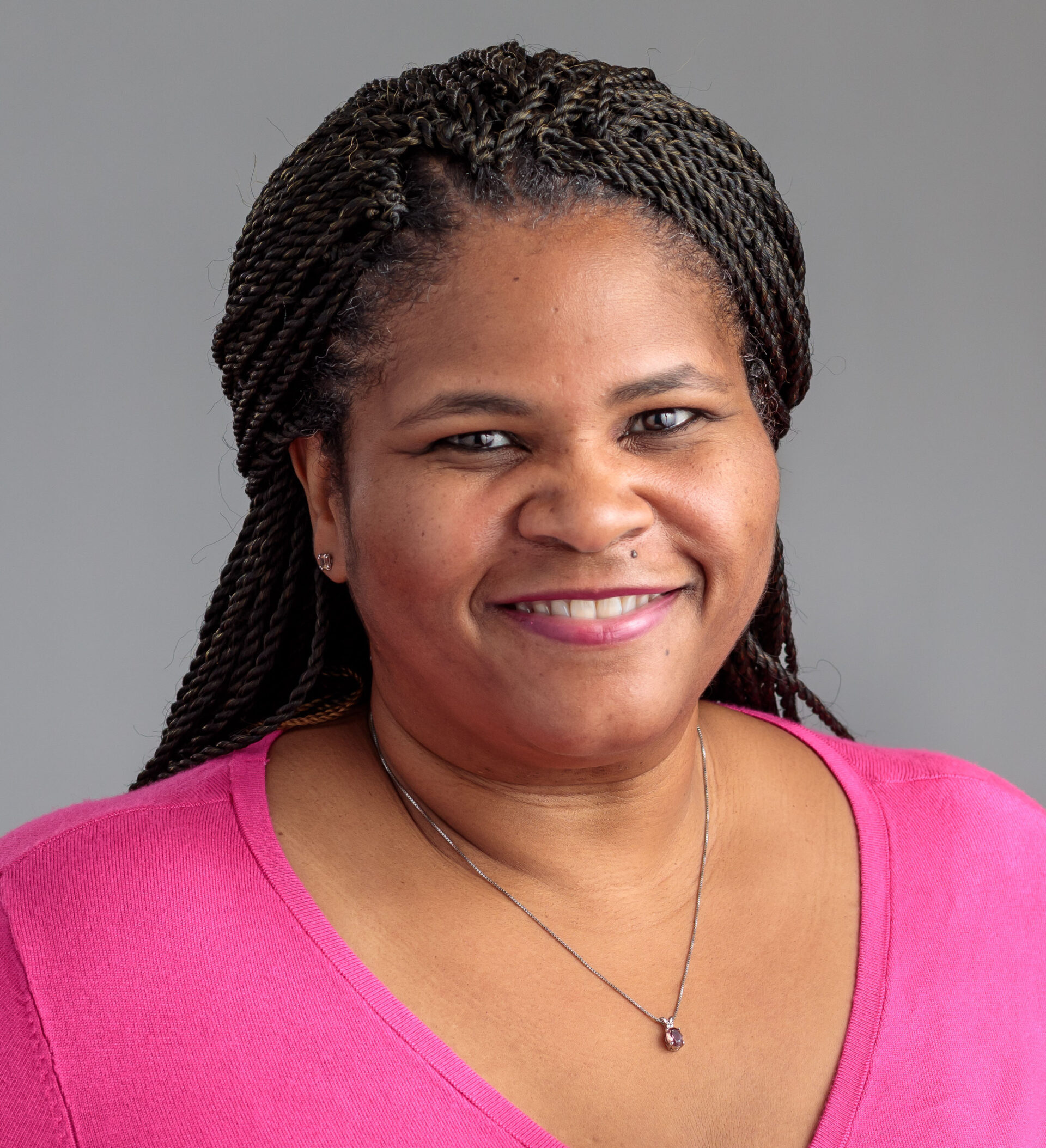 Professional headshot of Chandria Jones with long braided hair, wearing a bright pink top and a pendant necklace. The portrait is framed from the shoulders up against a light gray background
