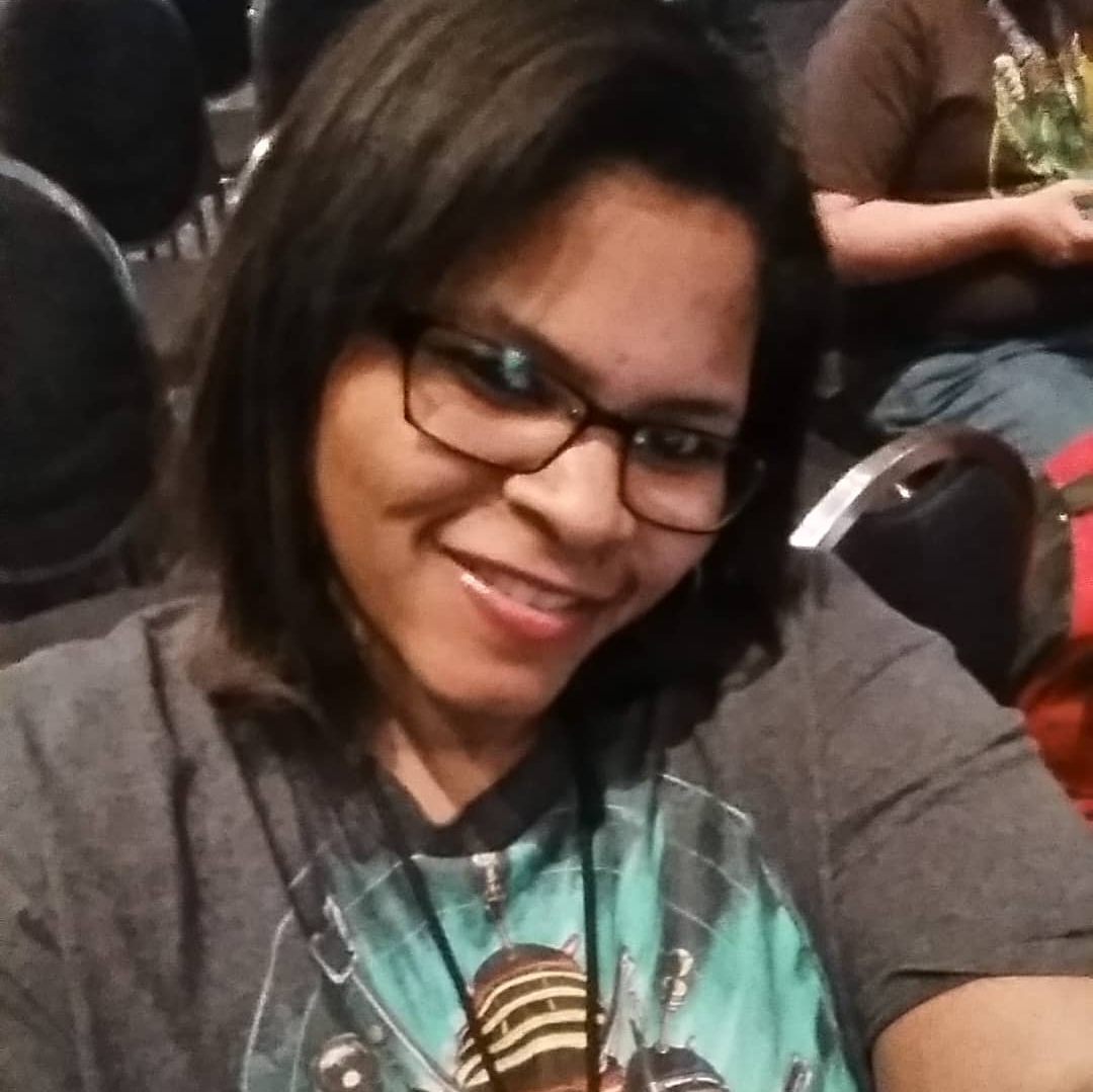 Close-up photo of Chandria Jones wearing rectangular glasses and a gray graphic T-shirt, seated indoors among rows of chairs at an event or gathering.