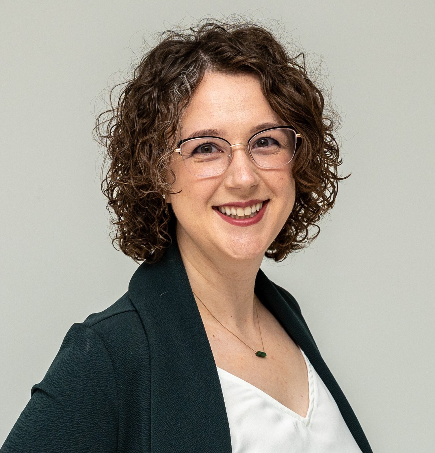 Professional headshot of Emily Royse with short, curly hair and glasses, wearing a dark blazer over a white blouse and a small necklace. The portrait is framed from the chest up against a light gray background.