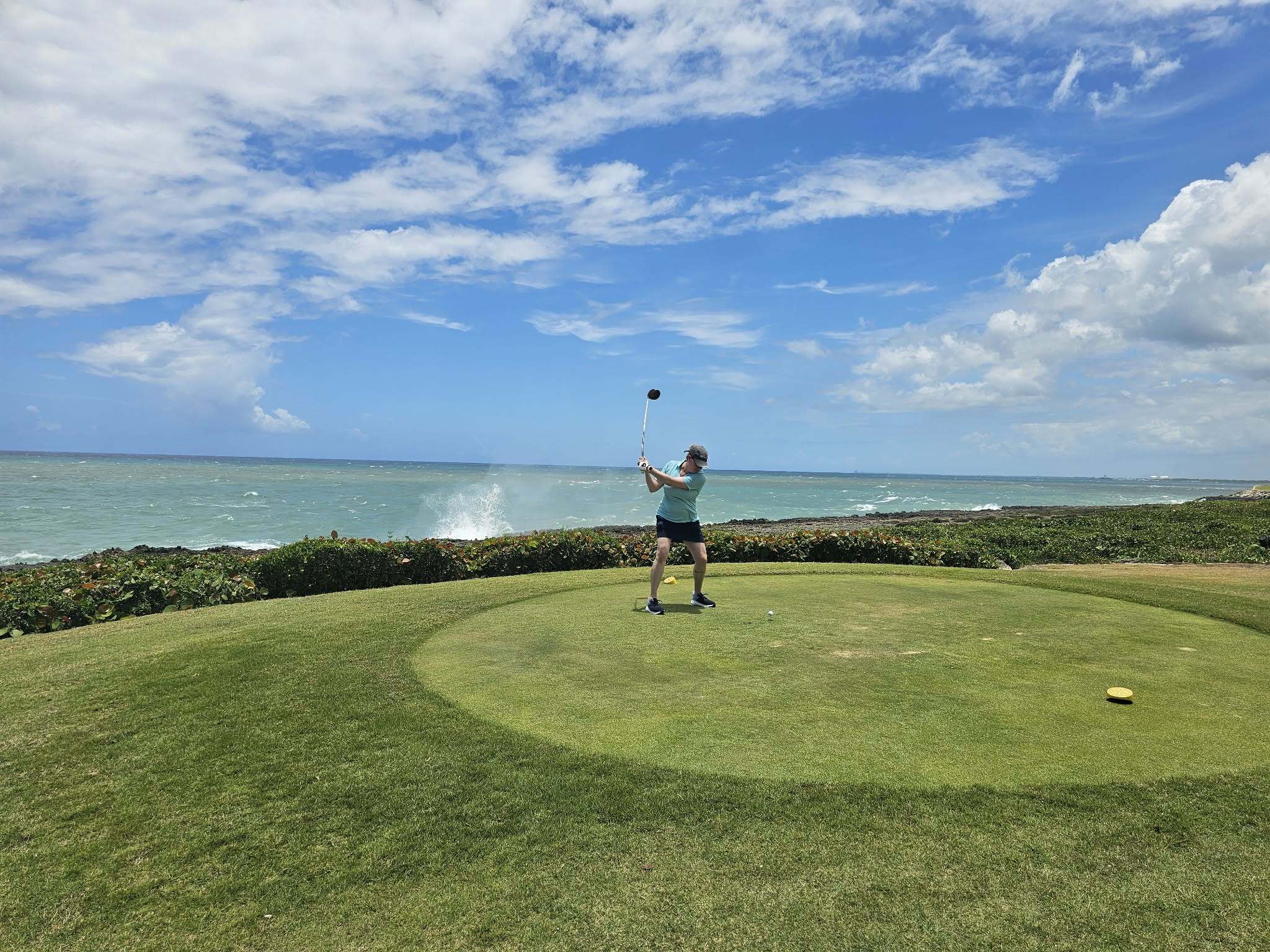 Jess Krim stands on a grassy tee box at a coastal golf course, swinging a golf club. The ocean stretches behind the course, with waves crashing against the shoreline and water spray visible in the distance. A bright blue sky filled with large white clouds spans the background, creating a scenic seaside setting.