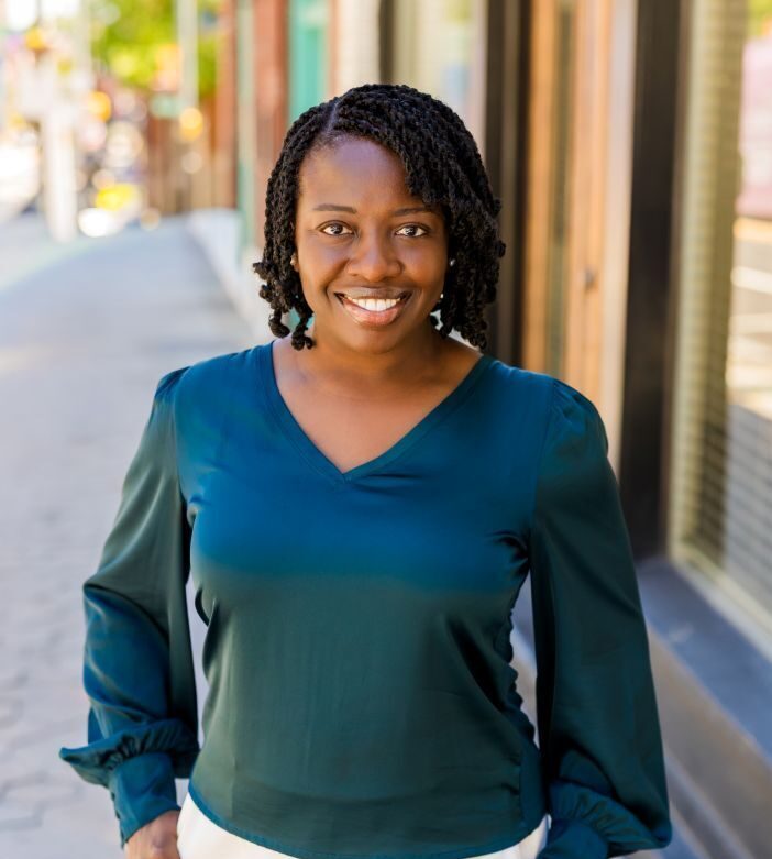 Professional outdoor portrait of a Linda Animashaun standing on a downtown sidewalk, wearing a teal blouse and white pants, with storefronts blurred in the background.