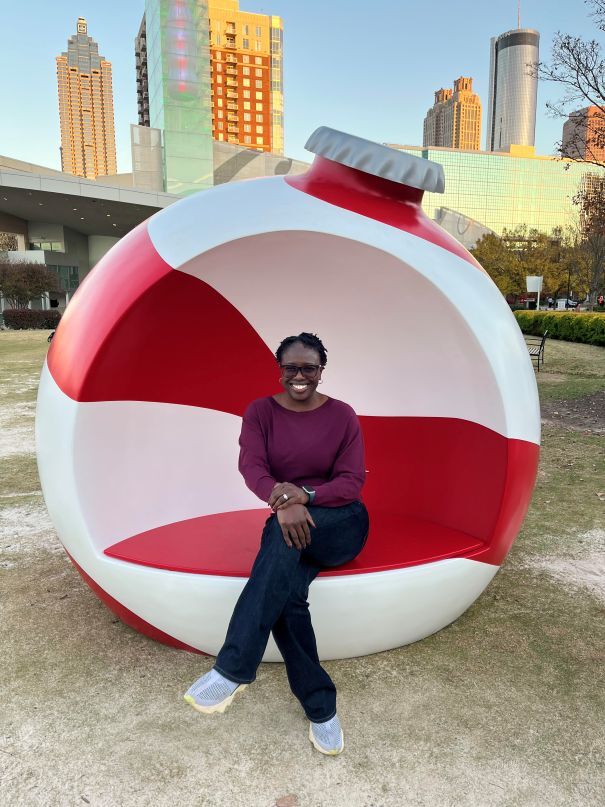 Linda Animashaun seated inside a giant red-and-white ornament decoration in a city park with skyscrapers in the background
