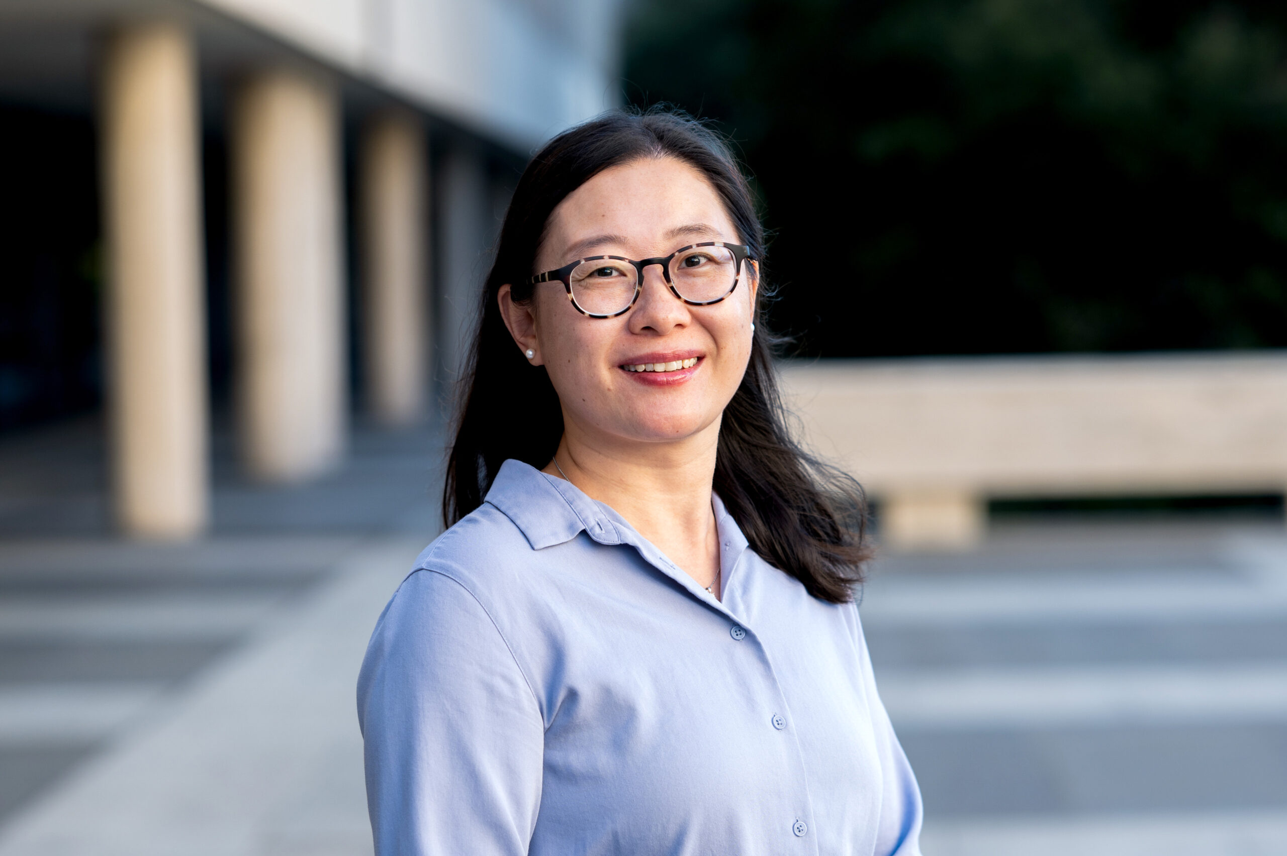 A professional outdoor headshot of Zhina Shen wearing glasses and a light blue collared blouse, standing in front of a modern building with columns