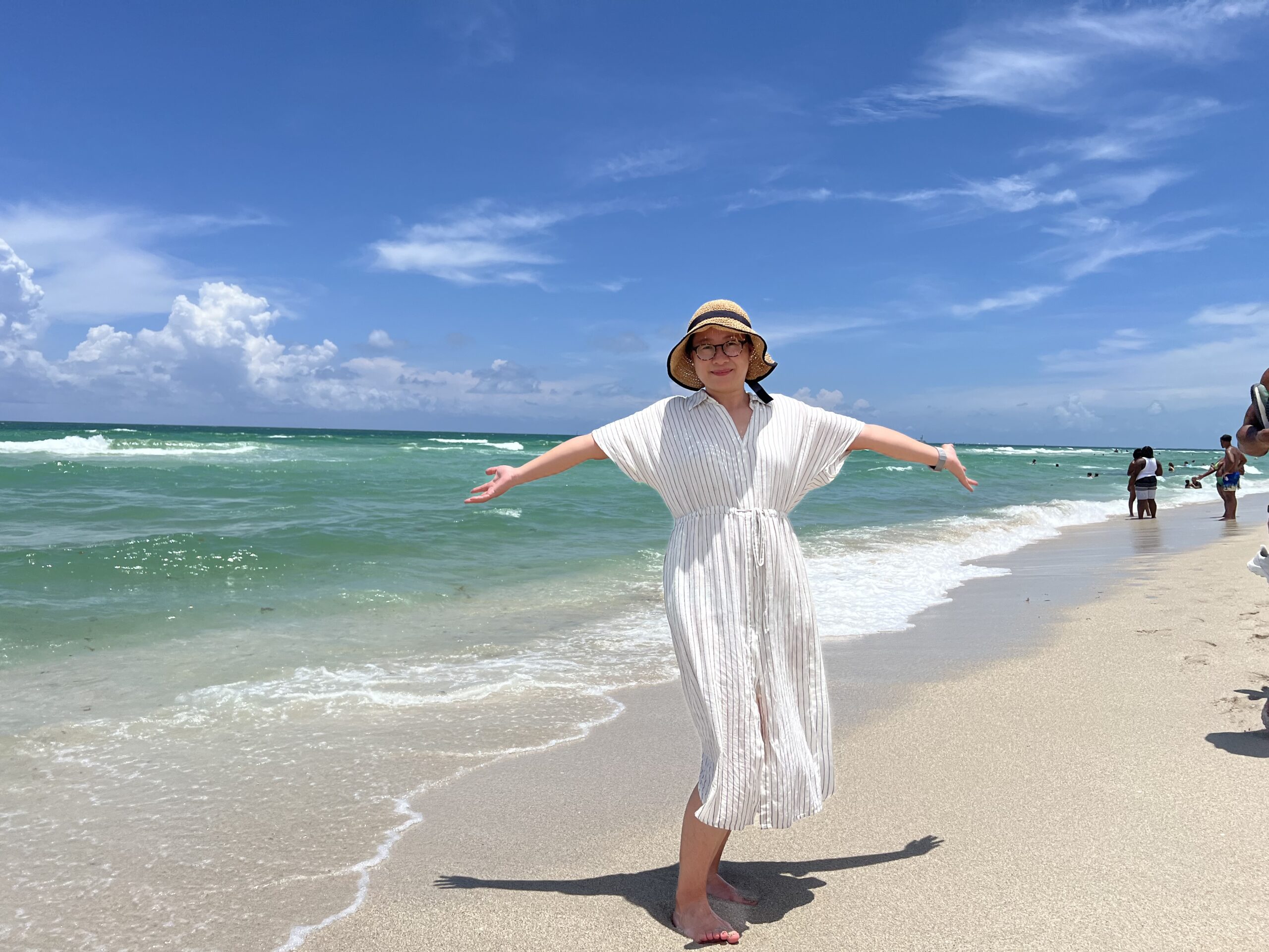 Zhina Shen wearing a wide-brimmed sun hat, glasses, and a light-colored striped beach cover-up stands barefoot on a sandy shoreline with arms outstretched. Turquoise ocean waves roll onto the beach under a bright blue sky with scattered white clouds.