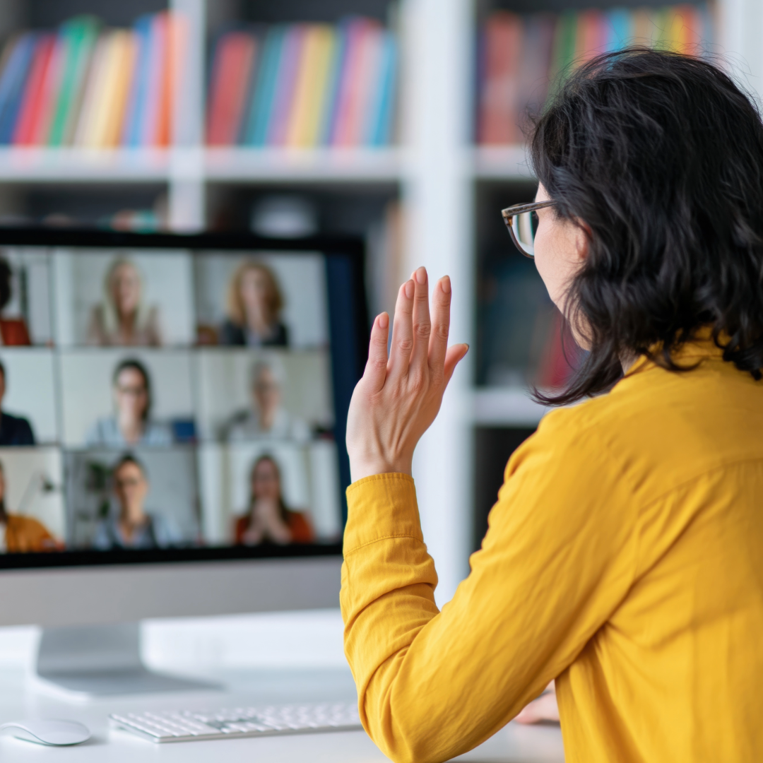 Woman in a gold shirt wavig at a computer screen with multiple people on it in a virtual meeting
