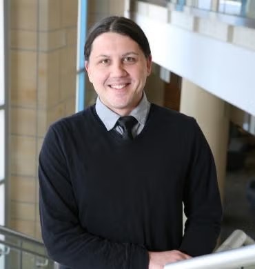 Professional head-and-shoulders portrait of Justin Sullivan standing indoors near a railing in a modern building with large windows and architectural columns. Justin is wearing a dark sweater over a collared shirt and tie, facing the camera. The background is softly blurred, showing an open atrium and upper-level walkway.