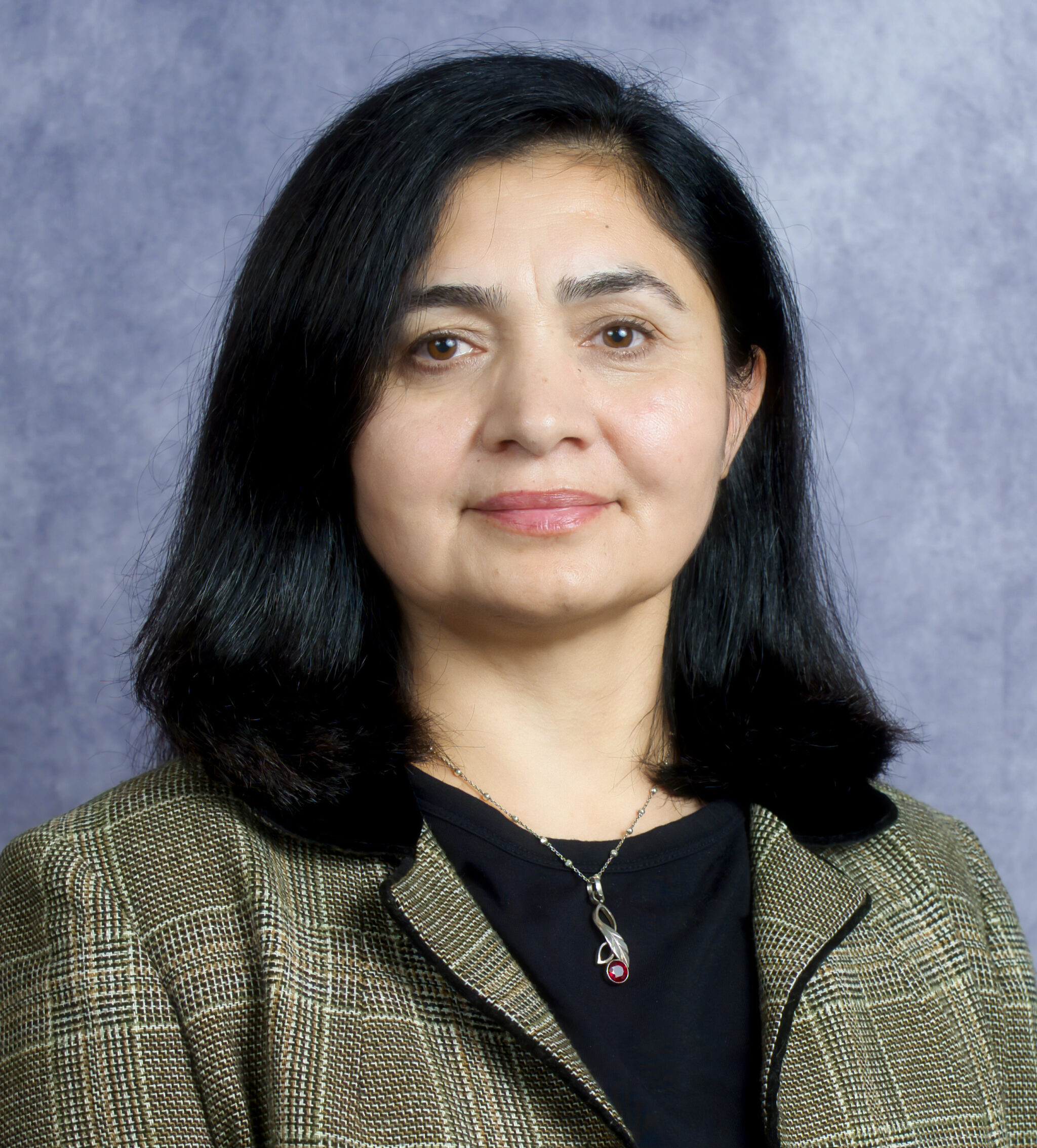 Professional headshot of Nahida Begum with shoulder-length dark hair wearing a patterned blazer over a dark top and a pendant necklace, photographed against a mottled blue background.